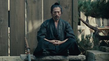 Movie still from “Westworld” (2016), created by Lisa Joy – A man sitting on a wall in a black robe; Medium shot, High angle