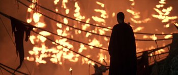 Movie still from “What Dreams May Come” (1998), directed by Vincent Ward – A man standing in front of a fire with wires behind him; Extreme Wide shot, Low angle