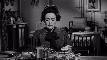 Movie still from “What Ever Happened to Baby Jane?” (1962), directed by Robert Aldrich – A woman sitting at a table with a plate in front of her face; Medium shot, High angle