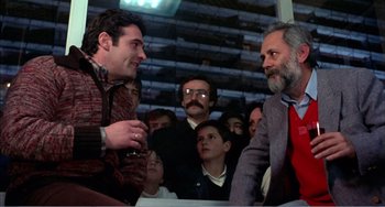 Movie still from “What Have I Done to Deserve This?” (1984), directed by Pedro Almodóvar – Two men talking to each other in front of a group of people; Medium shot, Over the shoulder angle