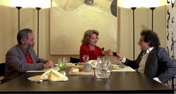 Movie still from “What Have I Done to Deserve This?” (1984), directed by Pedro Almodóvar – An older woman sitting at a dinner table with two other people; Medium shot, Over the shoulder angle