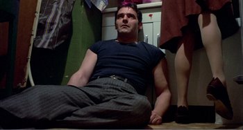 Movie still from “What Have I Done to Deserve This?” (1984), directed by Pedro Almodóvar – A man sitting on the ground in a room; Medium shot, Low angle