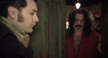 Movie still from “What We Do in the Shadows” (2014), directed by Taika Waititi – A man with long black hair standing next to another man with long black hair; Medium shot, Over the shoulder angle