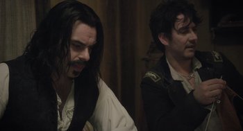 Movie still from “What We Do in the Shadows” (2014), directed by Taika Waititi – A man with long black hair and a beard; Medium shot, Over the shoulder angle
