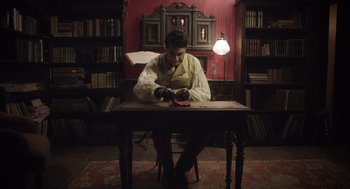 Movie still from “What We Do in the Shadows” (2014), directed by Taika Waititi – A man sitting at a table in front of a book case; Medium shot, High angle