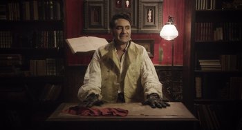 Movie still from “What We Do in the Shadows” (2014), directed by Taika Waititi – A man wearing gloves sitting at a table; Medium shot, Low angle