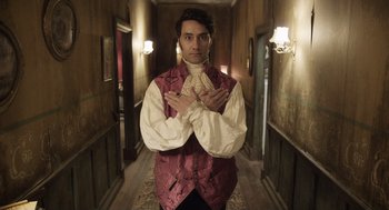 Movie still from “What We Do in the Shadows” (2014), directed by Taika Waititi – A man in a red vest standing in a hallway with his hands crossed; Medium shot, Overhead angle