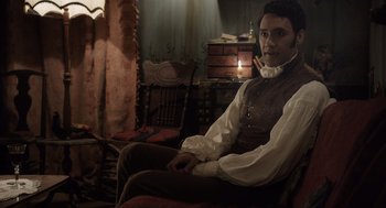 Movie still from “What We Do in the Shadows” (2014), directed by Taika Waititi – A man sitting on a chair in a room; Medium shot, Over the shoulder angle