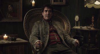 Movie still from “What We Do in the Shadows” (2014), directed by Taika Waititi – A man sitting in a wicker chair in a room; Medium shot, Low angle