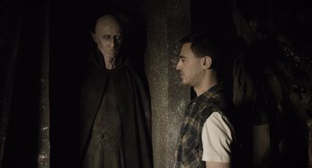 Movie still from “What We Do in the Shadows” (2014), directed by Taika Waititi – A man standing next to another man in a dark room; Medium shot, Over the shoulder angle