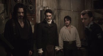 Movie still from “What We Do in the Shadows” (2014), directed by Taika Waititi – A man standing next to another man in a dark room; Medium shot, Low angle