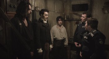 Movie still from “What We Do in the Shadows” (2014), directed by Taika Waititi – A group of men standing next to each other in a room; Medium shot, Over the shoulder angle