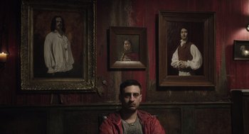 Movie still from “What We Do in the Shadows” (2014), directed by Taika Waititi – A man sitting in front of a wall with paintings on the wall; Medium shot, Low angle