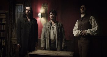 Movie still from “What We Do in the Shadows” (2014), directed by Taika Waititi – A group of men standing next to each other in front of a table; Medium shot, Low angle