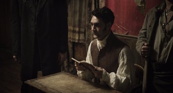 Movie still from “What We Do in the Shadows” (2014), directed by Taika Waititi – A man sitting at a table holding an open book; Medium shot, Over the shoulder angle