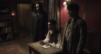Movie still from “What We Do in the Shadows” (2014), directed by Taika Waititi – A man sitting at a table with two other men standing next to him; Medium shot, Over the shoulder angle