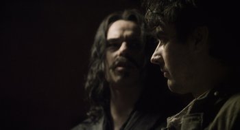 Movie still from “What We Do in the Shadows” (2014), directed by Taika Waititi – A man with long black hair standing next to another man with long black hair; Close Up shot, Over the shoulder angle