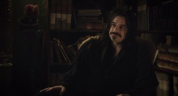Movie still from “What We Do in the Shadows” (2014), directed by Taika Waititi – A man with long hair sitting in front of a book shelf; Medium shot, Over the shoulder angle
