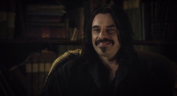 Movie still from “What We Do in the Shadows” (2014), directed by Taika Waititi – A man with long black hair sitting in a chair; Close Up shot, Over the shoulder angle