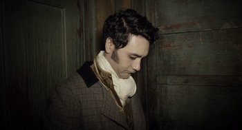 Movie still from “What We Do in the Shadows” (2014), directed by Taika Waititi – A man wearing a suit and tie standing next to a door; Close Up shot, High angle