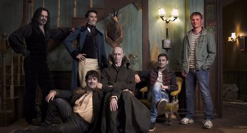 Movie still from “What We Do in the Shadows” (2014), directed by Taika Waititi – A group of men sitting on a couch in front of a painting; Wide shot, Low angle