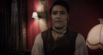 Movie still from “What We Do in the Shadows” (2014), directed by Taika Waititi – A man wearing a vest and a neck tie in front of a lamp; Close Up shot, Over the shoulder angle