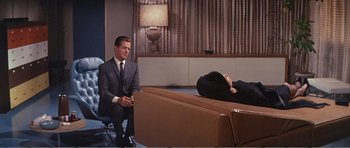 Movie still from “What a Way to Go!” (1964), directed by J. Lee Thompson – A man sitting on a couch next to a woman laying on the ground; Wide shot, Over the shoulder angle