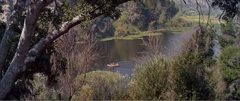 Movie still from “What a Way to Go!” (1964), directed by J. Lee Thompson – A person in a boat on a lake surrounded by trees; Extreme Wide shot, High angle