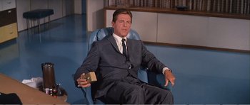 Movie still from “What a Way to Go!” (1964), directed by J. Lee Thompson – A man in a suit and tie sitting in a chair; Medium shot, Low angle