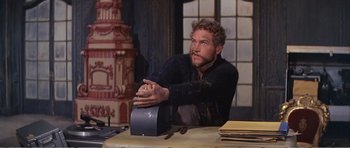 Movie still from “What a Way to Go!” (1964), directed by J. Lee Thompson – A man sitting at a table with his arms crossed; Medium shot, Low angle