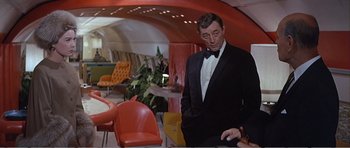 Movie still from “What a Way to Go!” (1964), directed by J. Lee Thompson – A man in a tuxedo standing next to a table in a room; Medium shot, Over the shoulder angle