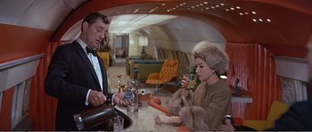 Movie still from “What a Way to Go!” (1964), directed by J. Lee Thompson – A man and a woman sitting at a table in an airplane; Medium shot, Over the shoulder angle