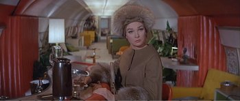 Movie still from “What a Way to Go!” (1964), directed by J. Lee Thompson – A woman in a fur hat sitting at a table in a room; Medium shot, Low angle