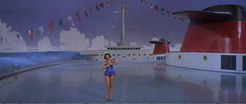 Movie still from “What a Way to Go!” (1964), directed by J. Lee Thompson – A woman in a blue dress standing in front of a ship; Extreme Wide shot, High angle