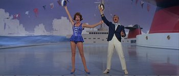 Movie still from “What a Way to Go!” (1964), directed by J. Lee Thompson – A man and a woman are holding their hands up in the air while standing in front of a ship; Wide shot, Low angle
