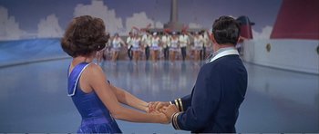 Movie still from “What a Way to Go!” (1964), directed by J. Lee Thompson – A couple of people holding hands in front of an audience; Medium shot, Over the shoulder angle