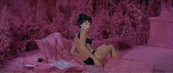 Movie still from “What a Way to Go!” (1964), directed by J. Lee Thompson – A woman sitting on a couch in front of pink bushes; Wide shot, High angle