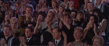 Movie still from “What a Way to Go!” (1964), directed by J. Lee Thompson – A group of people sitting in a crowd clapping; Medium shot, High angle