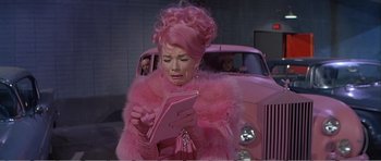 Movie still from “What a Way to Go!” (1964), directed by J. Lee Thompson – A woman in a pink dress is looking at a book; Medium shot, High angle
