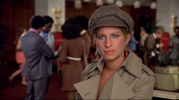 Movie still from “What's Up, Doc?” (1972), directed by Peter Bogdanovich – A woman wearing a hat and a trench coat in front of a group of people; Close Up shot, Over the shoulder angle