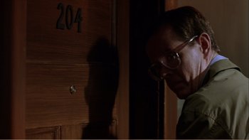 Movie still from “When a Man Falls” (2007), directed by Ryan Eslinger – A man with glasses standing in front of a wooden door; Close Up shot, Over the shoulder angle