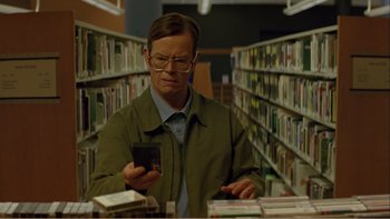 Movie still from “When a Man Falls” (2007), directed by Ryan Eslinger – A man in a library looking at a cell phone; Medium shot, Low angle