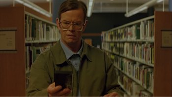 Movie still from “When a Man Falls” (2007), directed by Ryan Eslinger – A man holding a cell phone while standing in a library; Close Up shot, Low angle