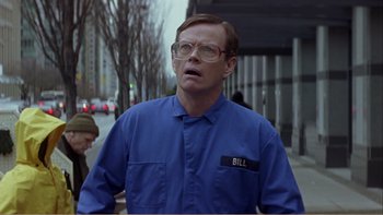 Movie still from “When a Man Falls” (2007), directed by Ryan Eslinger – A man in blue shirt and glasses on a sidewalk; Medium shot, Low angle