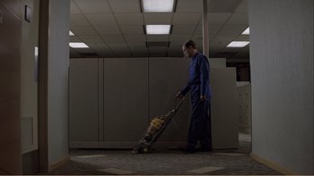 Movie still from “When a Man Falls” (2007), directed by Ryan Eslinger – A man in a blue suit is vacuuming in an office; Wide shot, Low angle