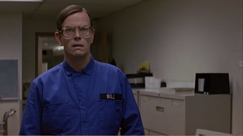 Movie still from “When a Man Falls” (2007), directed by Ryan Eslinger – A man wearing glasses and a blue shirt is standing in a room; Medium shot, Low angle