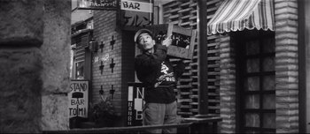 Movie still from “When a Woman Ascends the Stairs” (1960), directed by Mikio Naruse – A man holding up a record in front of a brick building; Medium shot, Low angle