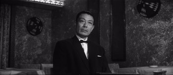 Movie still from “When a Woman Ascends the Stairs” (1960), directed by Mikio Naruse – A man in a suit standing in front of a wall; Medium shot, Low angle