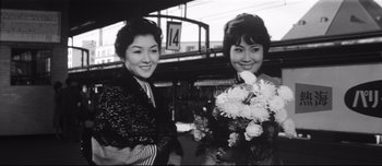 Movie still from “When a Woman Ascends the Stairs” (1960), directed by Mikio Naruse – Two young women standing next to each other holding flowers; Medium shot, Low angle