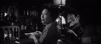 Movie still from “When a Woman Ascends the Stairs” (1960), directed by Mikio Naruse – A woman sitting in front of another woman holding a cell phone; Medium shot, Low angle